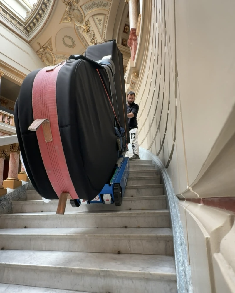 Pian cu coadă învelit, urcat pe o scară de marmură cu un cărucior cu șenile pentru piane