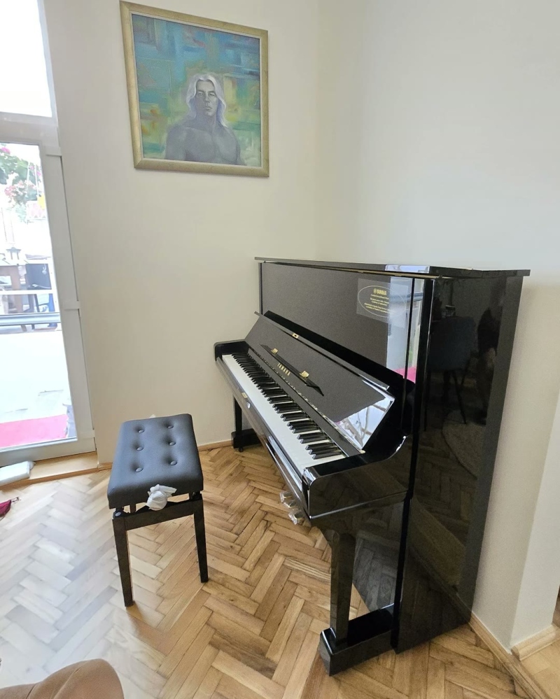 Pianină Yamaha neagră și o bancă de pian, într-o cameră cu parchet în spic
